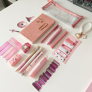 Pink stationery gift bundle with notebook, clear pencil pouch, pink pens, highlighters, sticky notes, correction tape, bookmarks, and bookish accessories arranged on a white background.