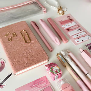 Close-up of a pink bookish stationery gift bundle with notebook, pens, pencil pouch, sticky notes, correction tape, bookmarks, and aesthetic desk accessories.