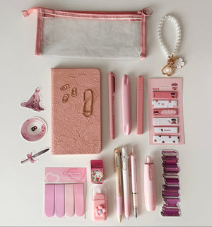 Flat lay of a pink stationery gift bundle with notebook, clear pencil pouch, pens, sticky notes, correction tape, bookmarks, keychain, and aesthetic desk accessories on a white background.