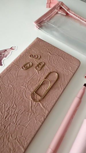 Close-up of a pink notebook with oversized gold paper clips, pink gel pen, and clear pencil pouch from a pink stationery gift bundle.