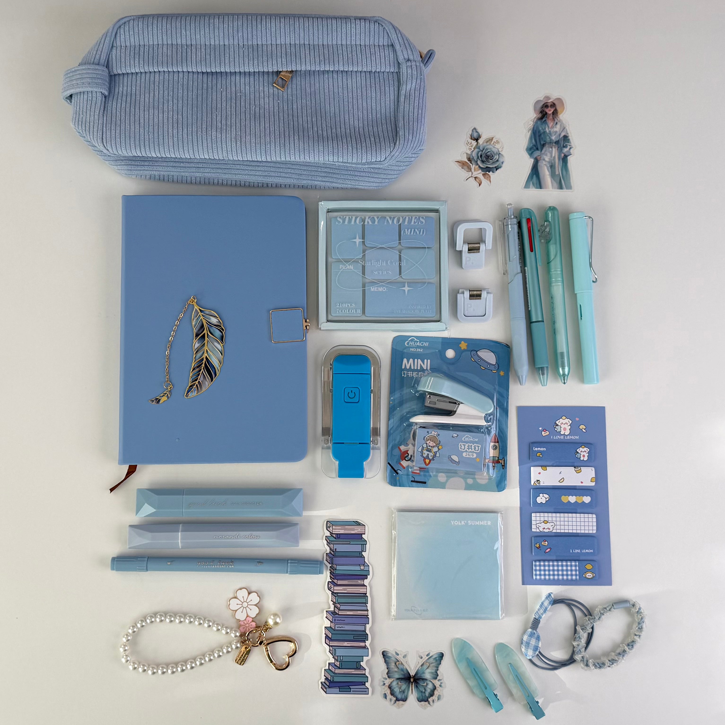 A flat lay of coordinated stationery and desk accessories in pastel blue tones on a white background, including a striped blue pencil case, notebook, pens, sticky notes, clips, a mini stapler, and decorative accessories.