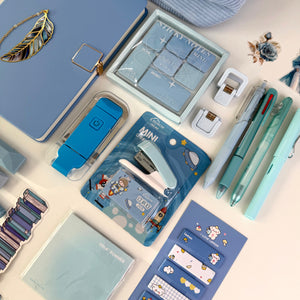 A styled flat-lay of pastel stationery items on a light beige surface, including a blue notebook, sticky notes, a mini stapler, pens, clips, and other desk accessories.