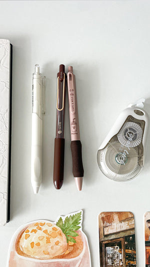 Close-up of a cream notebook, cream gel pen, two coffee-themed gel pens, correction tape, and coffee-inspired stationery from the Morning Coffee Stationery Gift Bundle.