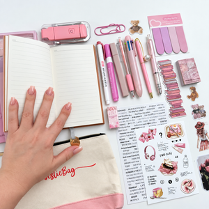 Open pink A6 notebook showing lined writing pages, surrounded by pastel pens, highlighters and decorative journaling supplies.