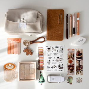 A styled flat-lay of various stationery and beauty accessories on a white surface with soft sunlight shadows. The collection includes a cream-colored fabric pouch, a brown textured notebook, pens, a mini stapler, stickers, nail files, and other desk accessories.