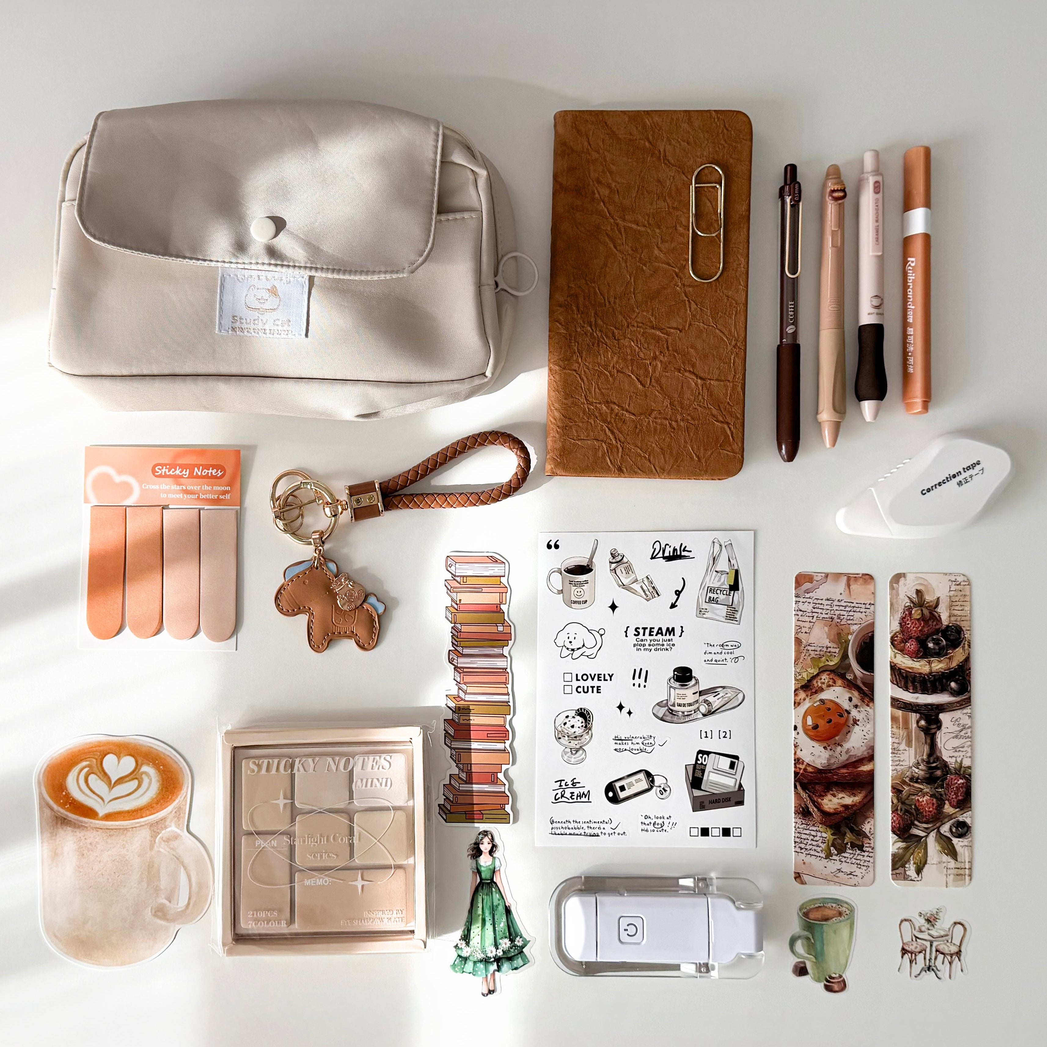 A styled flat-lay of various stationery and beauty accessories on a white surface with soft sunlight shadows. The collection includes a cream-colored fabric pouch, a brown textured notebook, pens, a mini stapler, stickers, nail files, and other desk accessories.