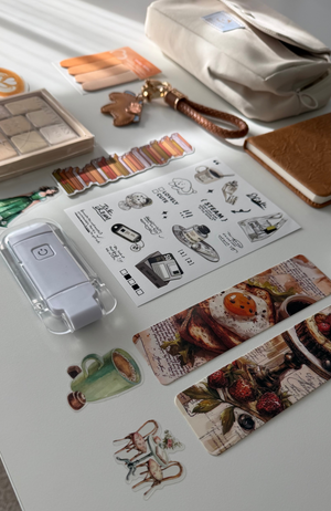 A styled flat-lay of various stationery and desk accessories on a white surface, including a clear plastic case with a white device, sticker sheets with illustrations and food prints, a cream pouch, and a notebook.