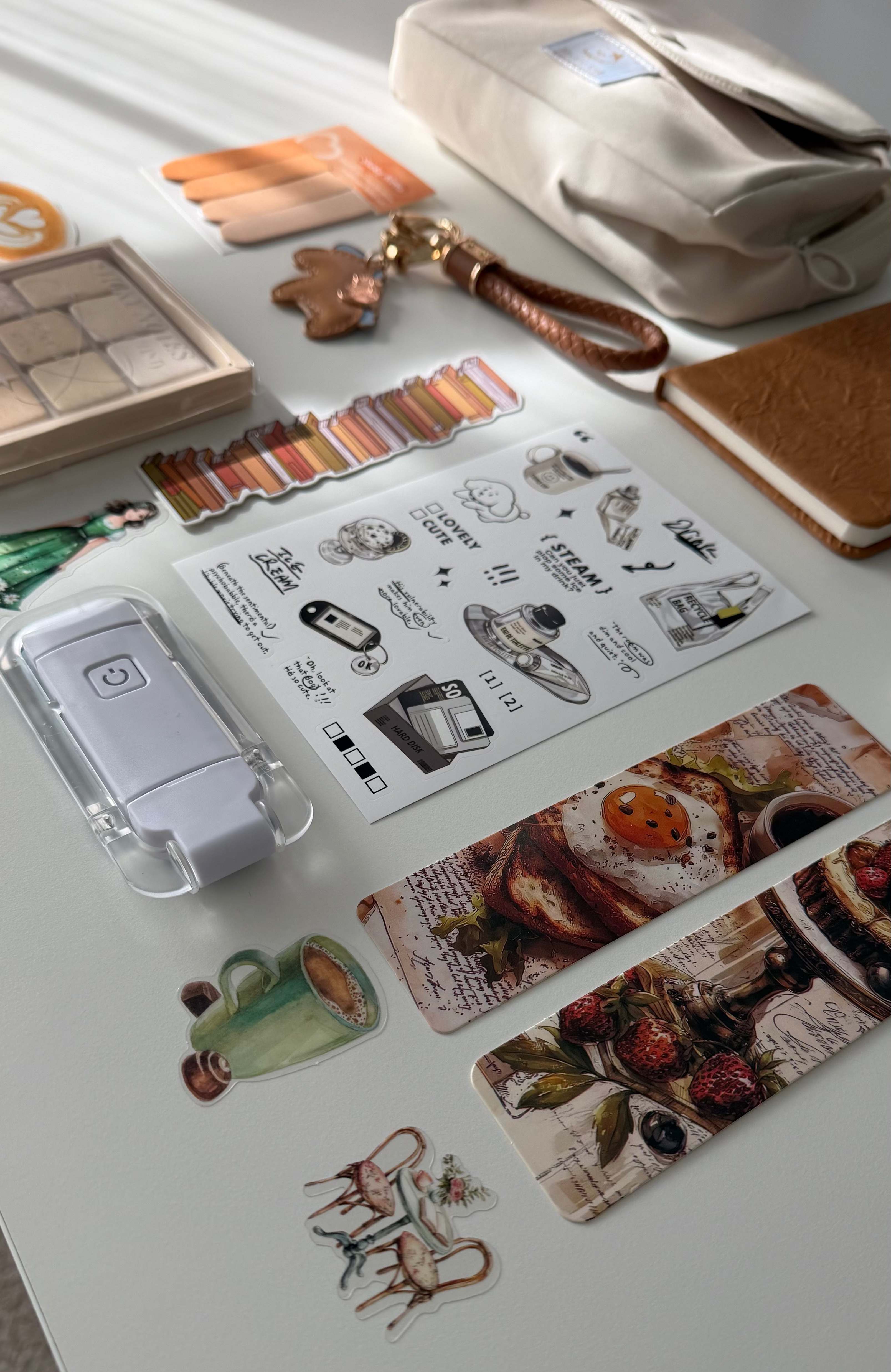 A styled flat-lay of various stationery and desk accessories on a white surface, including a clear plastic case with a white device, sticker sheets with illustrations and food prints, a cream pouch, and a notebook.