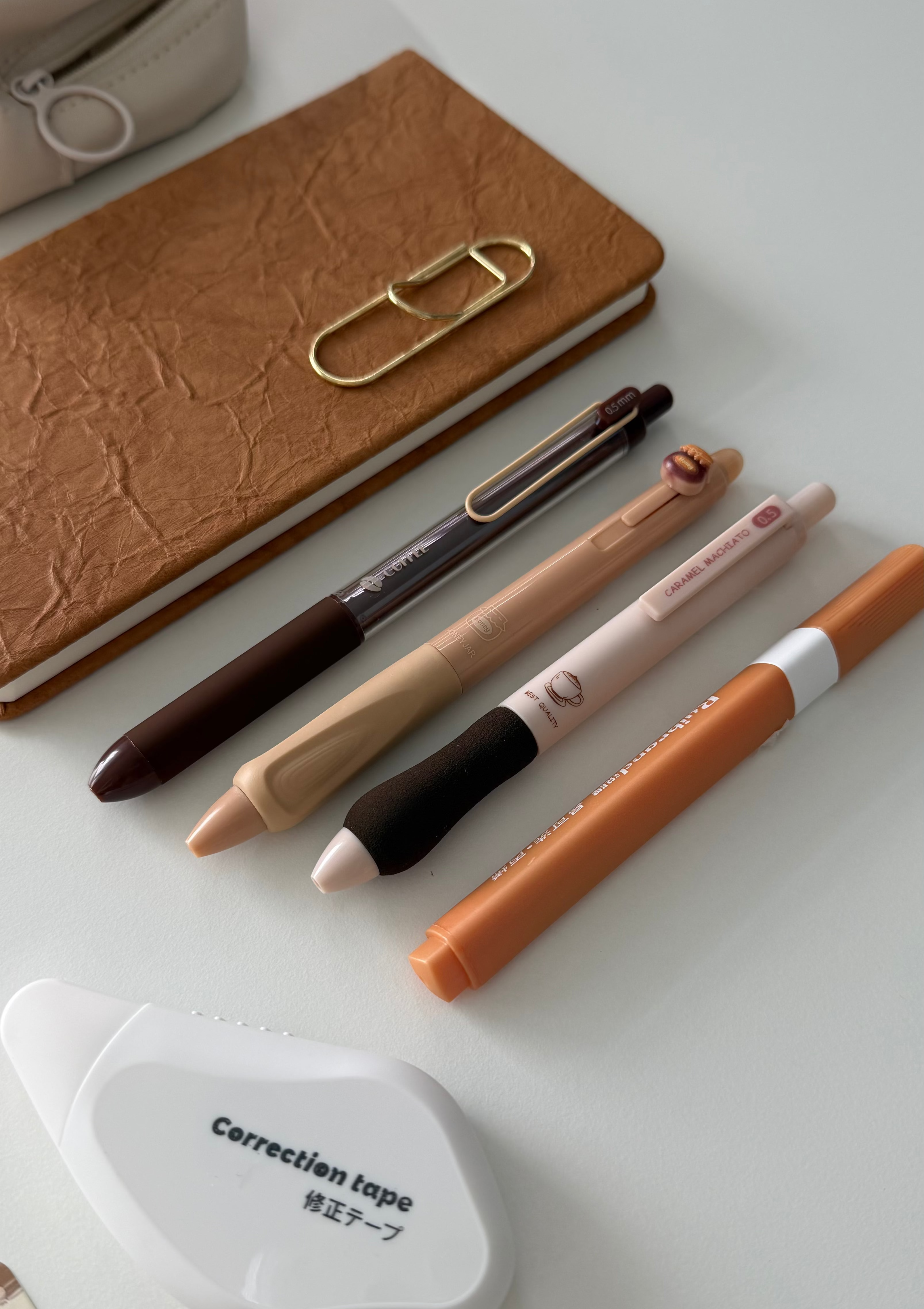 A styled flat-lay of stationery items on a light gray surface, including a brown textured notebook with a gold paperclip, several pens, a white correction tape dispenser, and part of a beige pencil case.