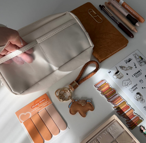 A hand holds a cream-colored pencil pouch on a white desk, surrounded by stationery, nail files, a compact powder, and a cup of latte art coffee.