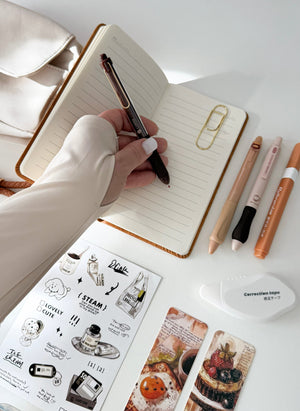 A top-down view of a desk with a hand writing in an open lined notebook using a black and silver pen. Surrounding the notebook are stationery items including a gold paperclip, three colored pens, a white correction tape dispenser, and two decorative bookmarks with food illustrations.