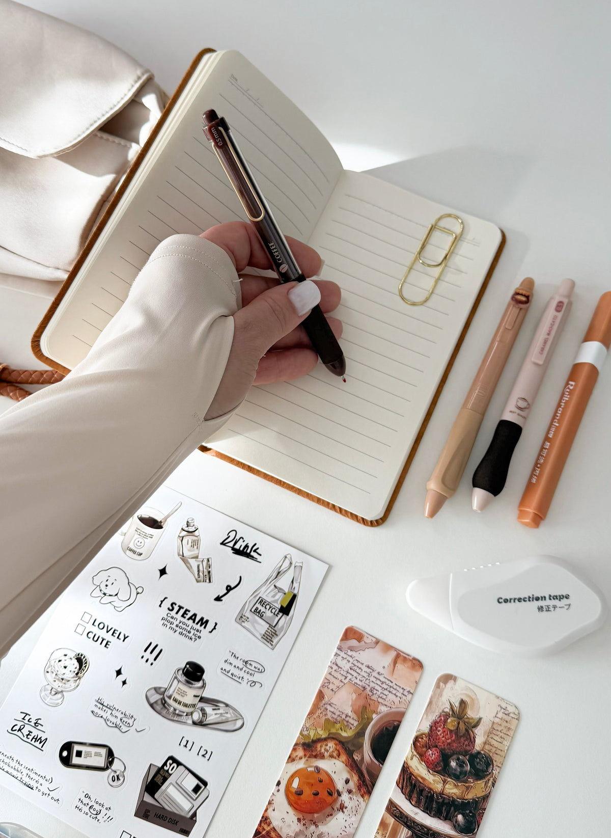A top-down view of a desk with a hand writing in an open lined notebook using a black and silver pen. Surrounding the notebook are stationery items including a gold paperclip, three colored pens, a white correction tape dispenser, and two decorative bookmarks with food illustrations.