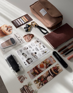 A styled flat-lay of a coffee-themed stationery and vanity set on a white table with natural light. The Gift stationery set includes a brown canvas pouch, a brown notebook, a black coffee mug, a black coffee bean keychain, a mini color palette, a sticker sheet with coffee illustrations, and various coffee-themed stickers.