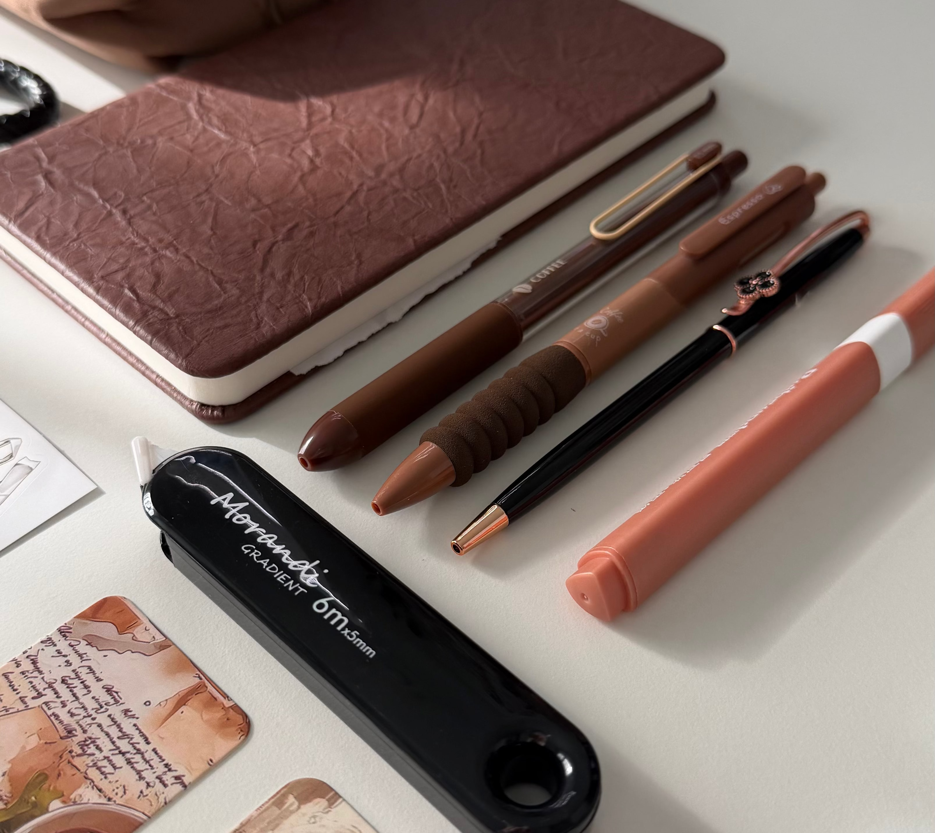 A black Magic Wand stylus pen with a white tip lies on a white desk next to a brown notebook, pens, a peach highlighter, and bookmarks.