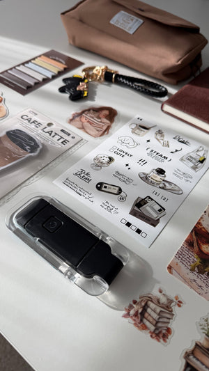 A styled flat-lay of stationery and accessories on a white surface, featuring a clear acrylic holder with a black flip phone, coffee-themed sticker sheets, a brown pouch, and other desk accessories.