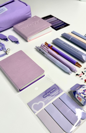 A collection of pastel purple stationery and desk accessories arranged on a white surface, including a zip pouch, notebooks, pens, sticky notes, and nail files.