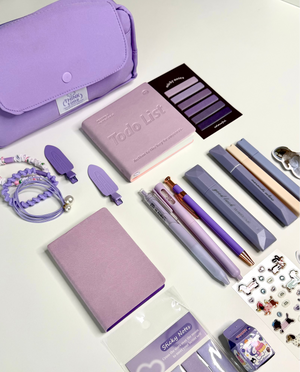 A coordinated stationery and accessory set in pastel purple and lavender tones arranged on a white tabletop. Visible items include a large lavender pouch, a pink To-do list notebook, two lavender notebooks, several purple and gray pens, a lavender bracelet, small accessories, and packaged sticky notes and stickers.