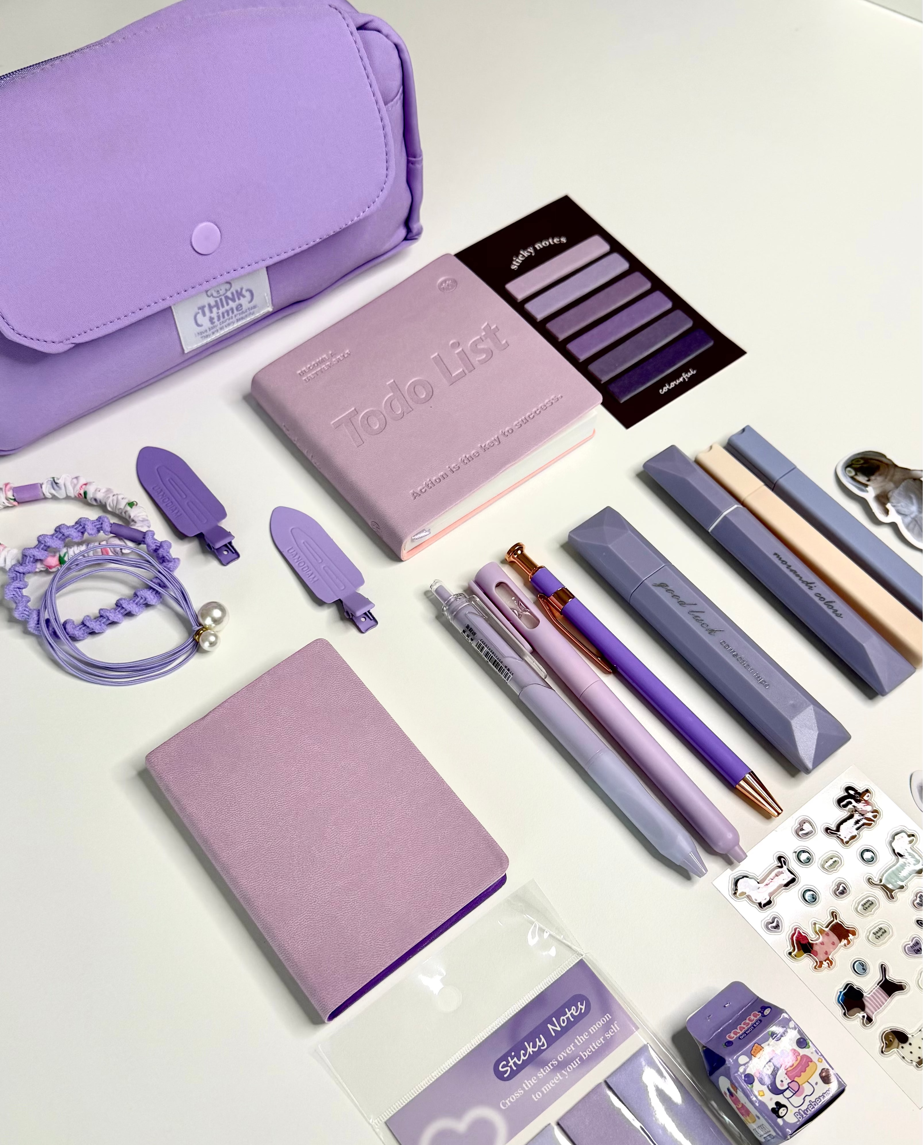 A coordinated stationery and accessory set in pastel purple and lavender tones arranged on a white tabletop. Visible items include a large lavender pouch, a pink To-do list notebook, two lavender notebooks, several purple and gray pens, a lavender bracelet, small accessories, and packaged sticky notes and stickers.