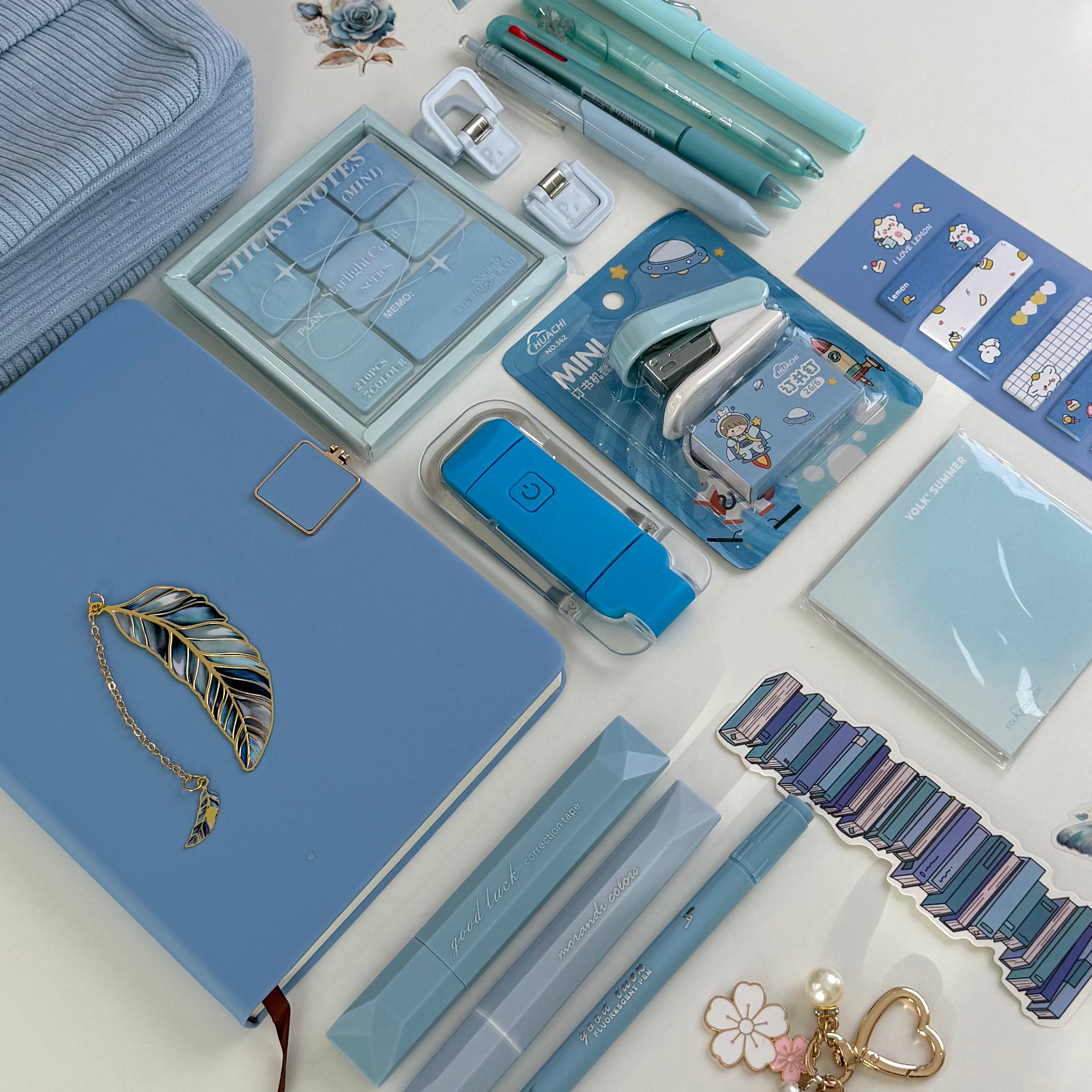 A flat-lay arrangement of pastel blue stationery and desk accessories on a white surface, including a notebook with a feather clasp, pens, a highlighter, a mini stapler, sticky notes, a wrist strap, and small stationery items.