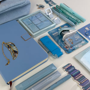 A styled flat-lay of stationery items on a white surface, including a light blue notebook with a gold feather bookmark, mint pens, a blue highlighter, a clear plastic organizer, a blue eraser, and other matching blue stationery accessories.