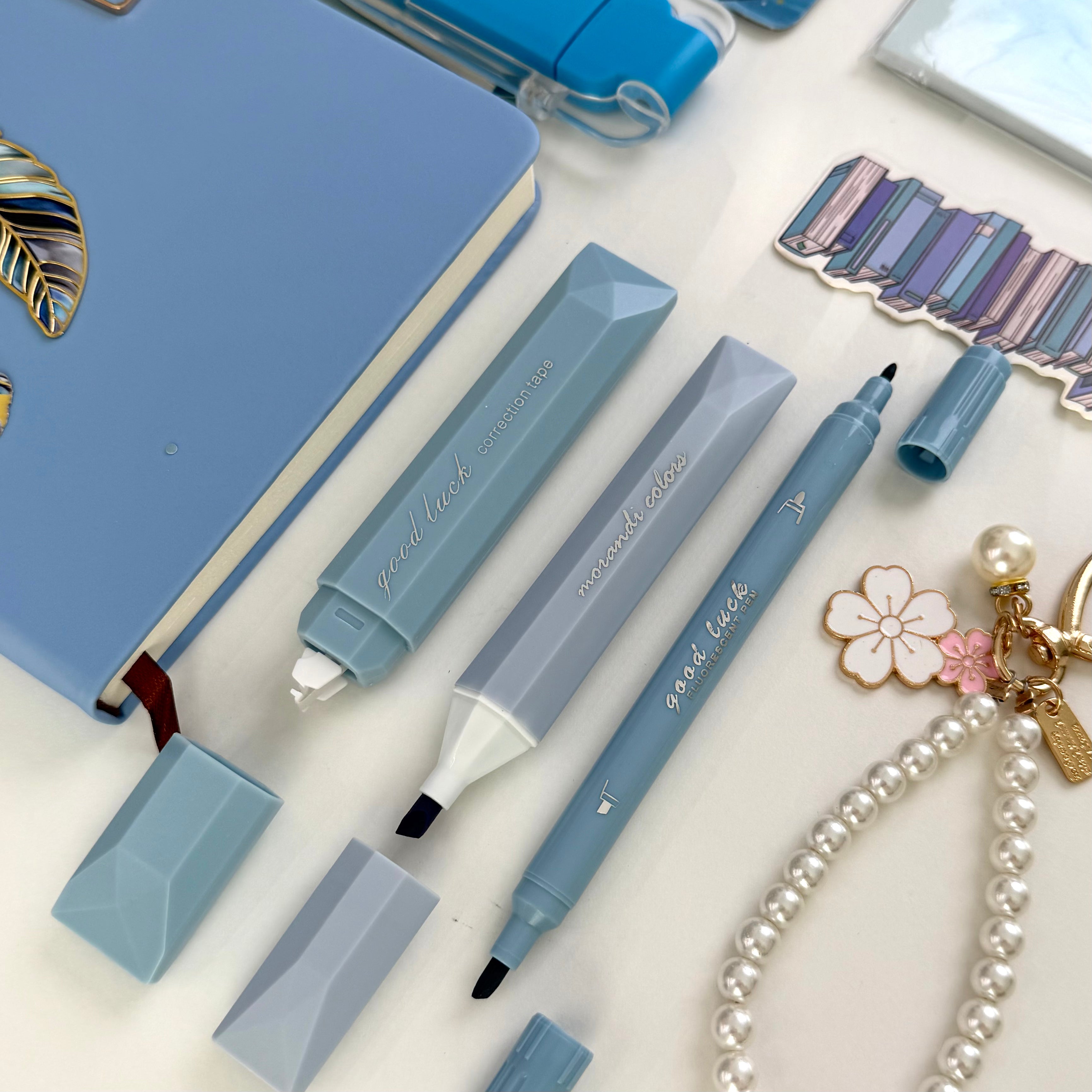 A styled flat-lay of light blue and gray faceted dual-tip highlighters on a white surface, alongside a blue notebook, sticker sheets, and a pearl flower keychain.